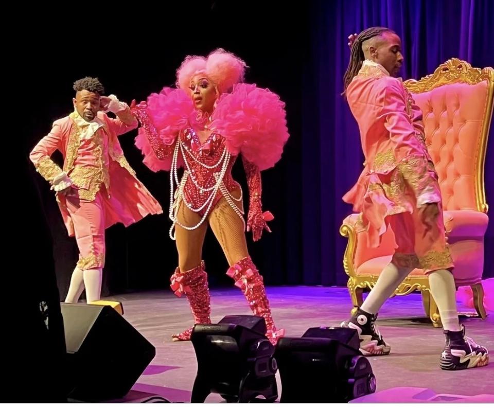 A drag queen with a pink wig, wearing sparkled pink gloves, boots, and top, with several pearl necklaces, stands onstage by a gilded pink throne-like chair, between two people wearing 18th century style clothing with converse