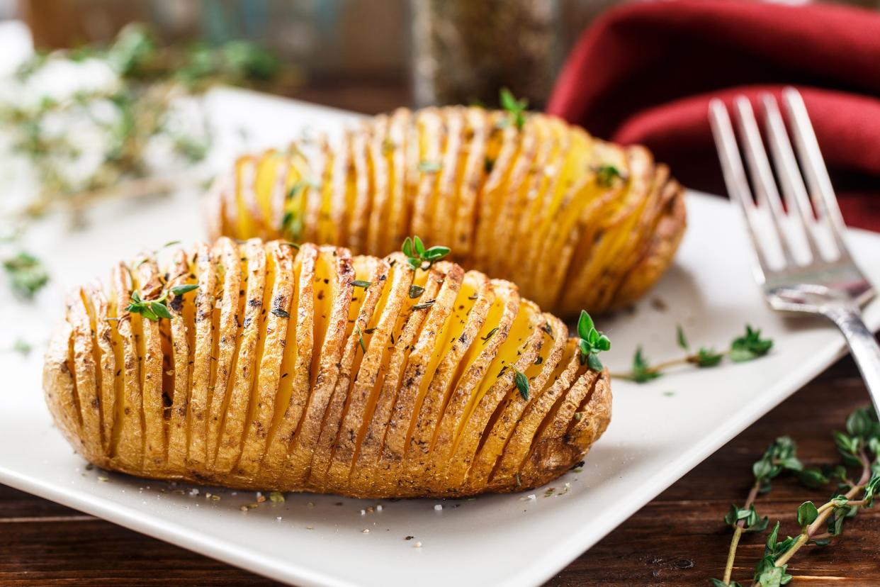 Two hasselback potatoes on a white plate with garnishes and a fork and a blurred background