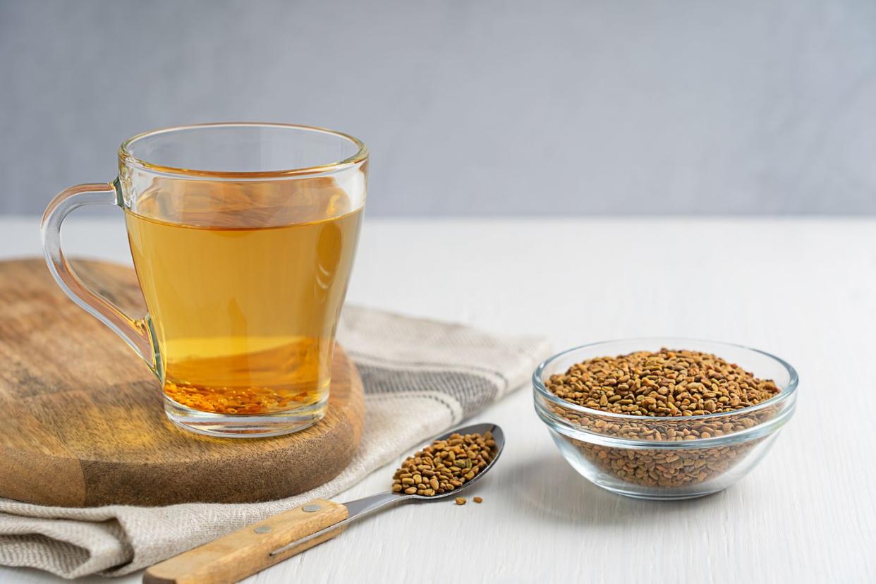helba golden tea drink made of fenugreek annual plant served in glass cup with bowl of seeds and spoon on white wooden table with textile used in medicine of egypt and as food ingredient horizontal