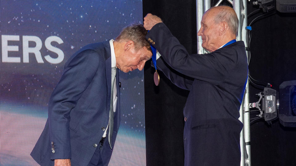 An older man pulls a coin around the neck of a man on stage