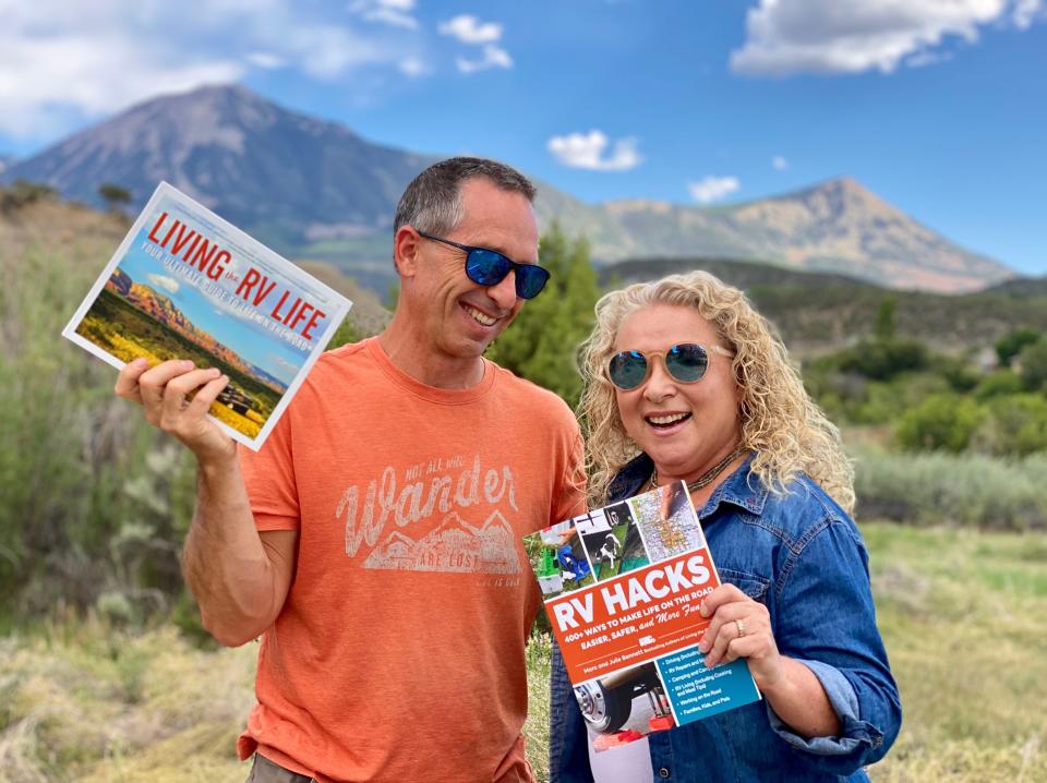 Marc and Julie Bennett of RVLove with RV Hacks and Living the RV Life books and mountains and blue skies in the background.