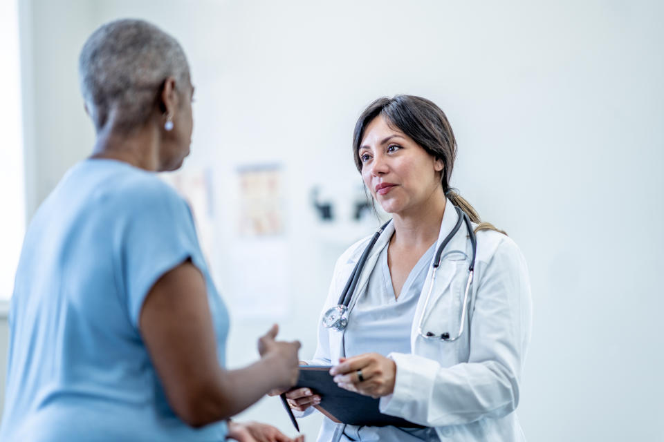 A senior woman of African decent, sits up on an exam table during a routine check-up with her doctor.  The doctor is seated in front of her and wearing scrubs as the two talk. Women have historically been ignored in the healthcare system. Here are expert tips on how to advocate for yourself at the doctor's. (Image via Getty)