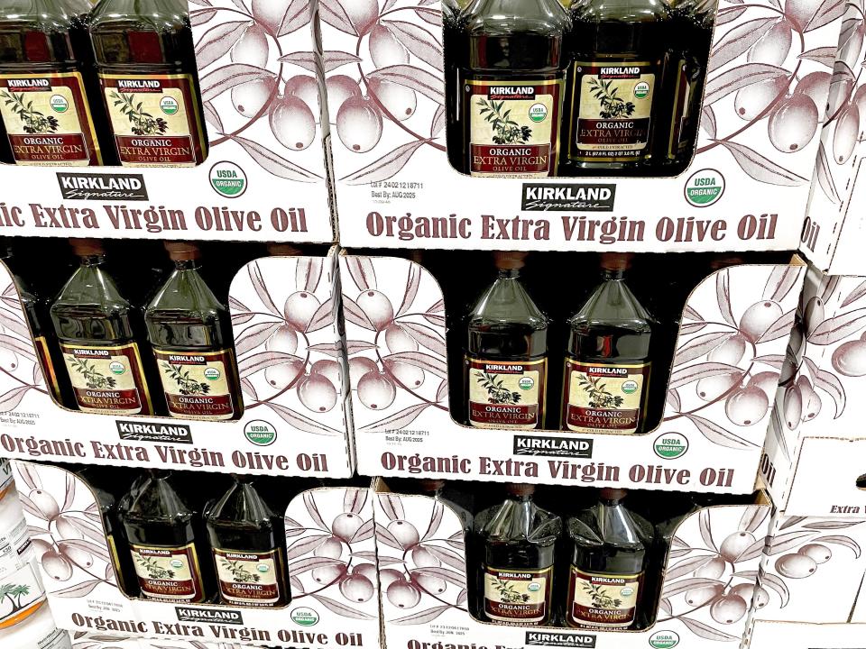 Bottles of Kirkland Signature olive oil with images of olive branches on the yellow label. The bottles are in white cardboard boxes with illustrations of olives on them