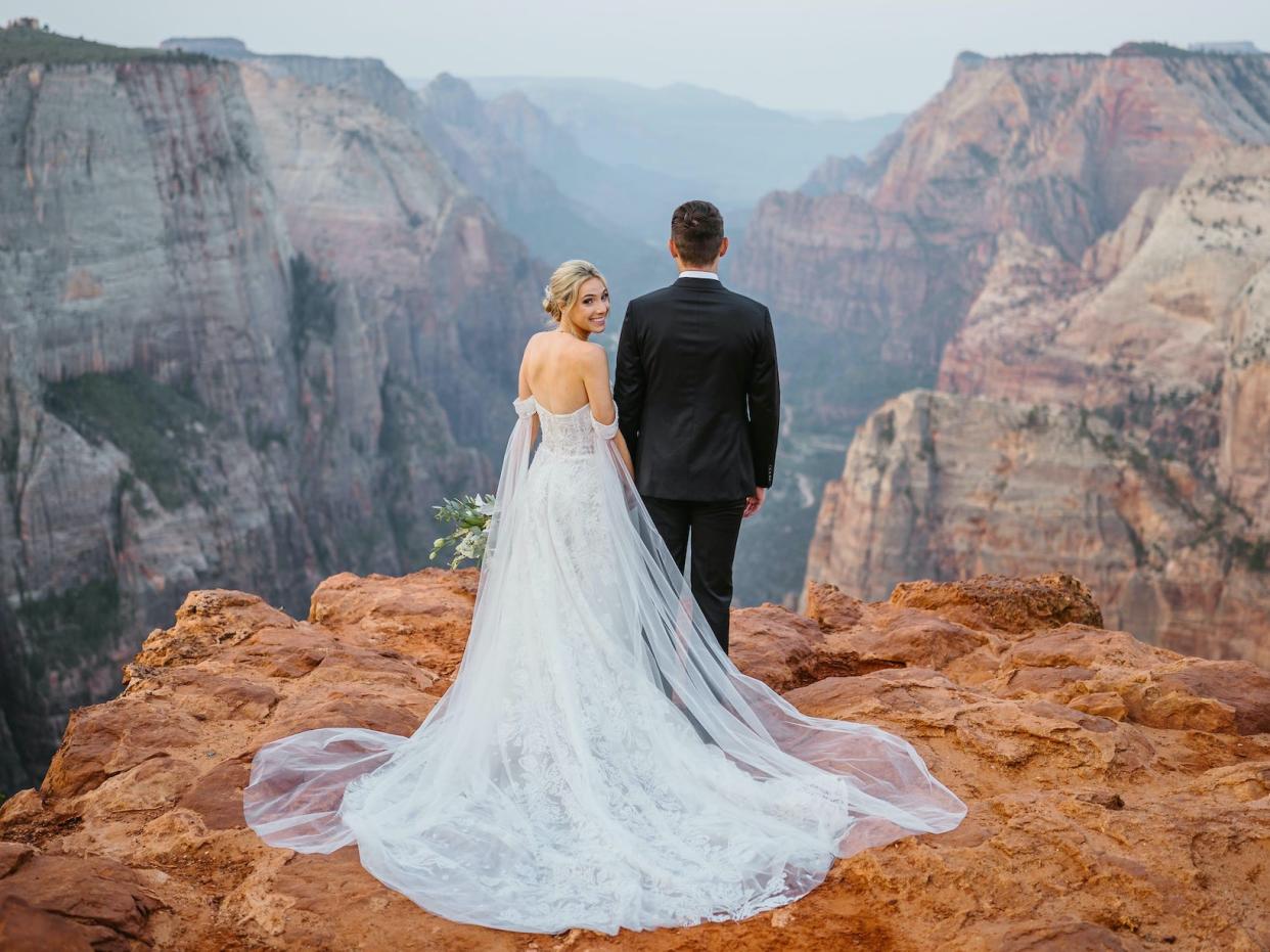 A bride looks over her shoulder while holding hands with her groom on top of a mountain.