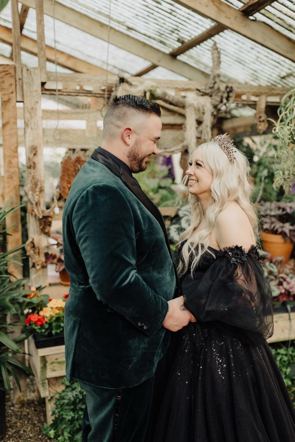 A bride and groom hold hands and look at each other in a greenhouse.