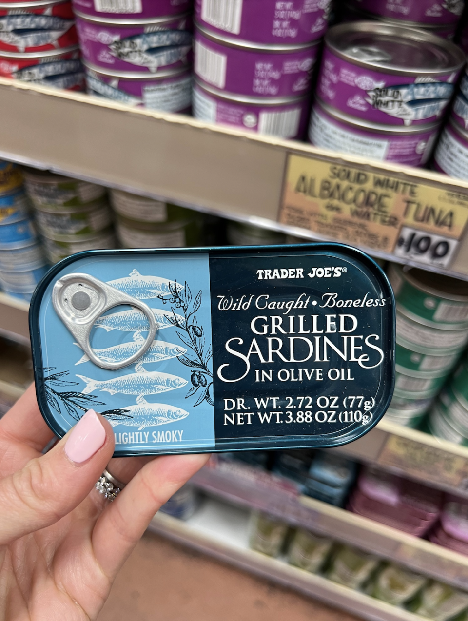 Hand holding a tin of Trader Joe's Wild Caught Boneless Grilled Sardines in Olive Oil, Lightly Smoky, in front of store shelves with various canned goods