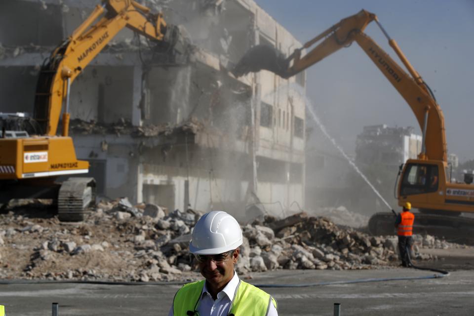 Greece's prime minister Kyriakos Mitsotakis wearing a helmet and plastic glasses stands at the old airport as bulldozers demolish an abandoned building in Athens, Friday, July 3, 2020. Mitsotakis inaugurated the start of construction work on a long-delayed major development project at the prime seaside site of the old Athens airport. The development of the 620-hectare (1,500-acre) Hellenikon site was a key element of the privatization drive that was part of Greece's international bailouts. (AP Photo/Thanassis Stavrakis)