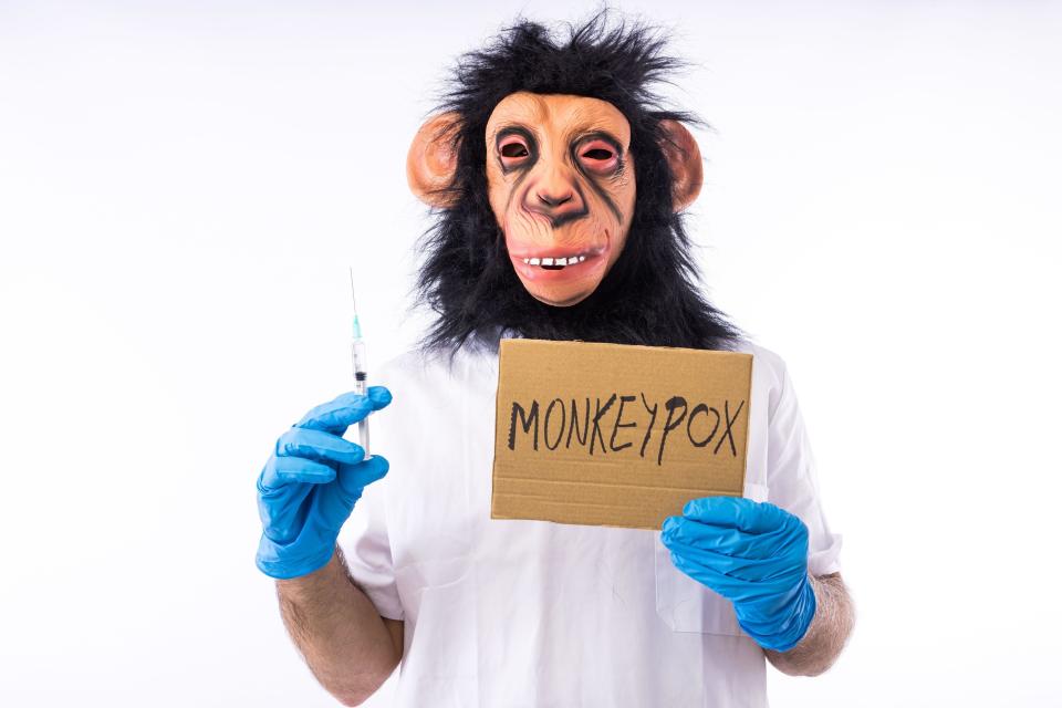 A man wearing a doctor's coat and gloves, holding a syringe and a monkeypox sign while wearing a monkey mask