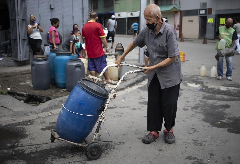 A man, wearing a protective face mask, pushes a dolly holding a container filled with water he collected from a street faucet, in Caracas, Venezuela, Saturday, June 20, 2020. Water service in Venezuela has gotten so bad that poor neighborhoods have started to rig private water systems or hand dig shallow wells. (AP Photo/Ariana Cubillos)