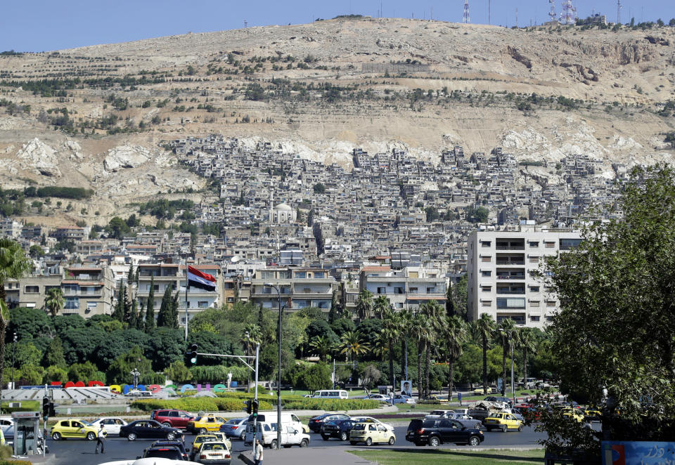 FILE - In this Aug. 13, 2018 photo taken through a bus window Syrians drive their cars in front of residential buildings in Damascus, Syria. A new Syrian law empowering the government to confiscate property is threatening to leave refugees stuck in Europe with no homes to return to. (AP Photo/Sergei Grits)