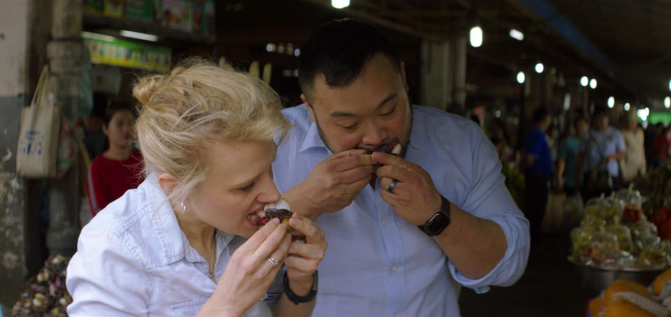 This image released by Netflix shows David Chang, right, and actress Kate McKinnon trying fresh mangosteen from a street vendor in Phnom Penh, Vietnam. in a scene from the Netflix series, "Breakfast, Lunch & Dinner." (Netflix via AP)