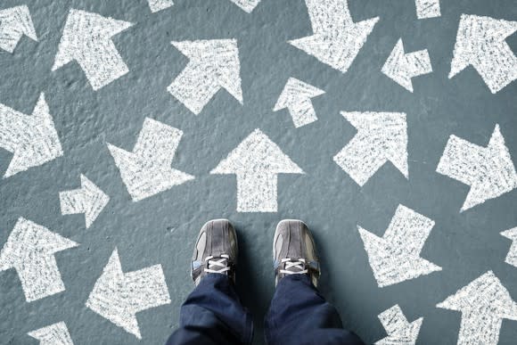 The point of view of someone looking down at their feet, and white arrows pointing in various directions are painted on the ground.