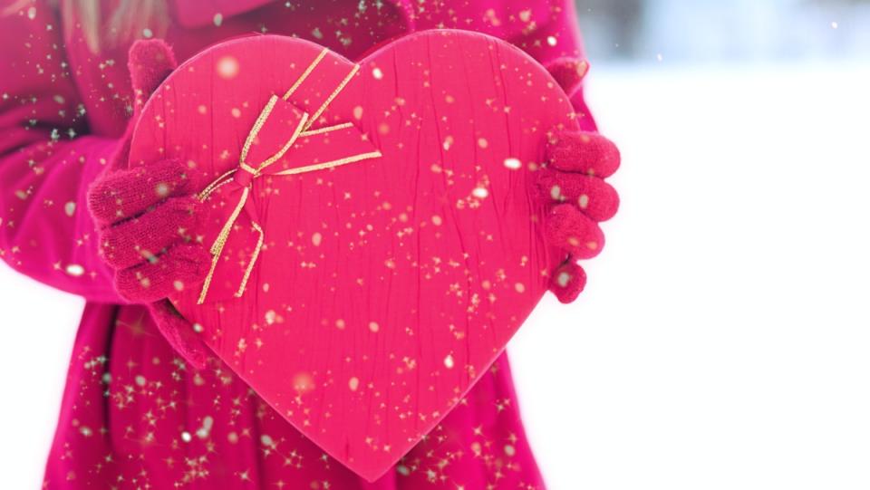 a woman wearing red coat and gloves stands in the snow holding a box of Valentine's Day chocolates