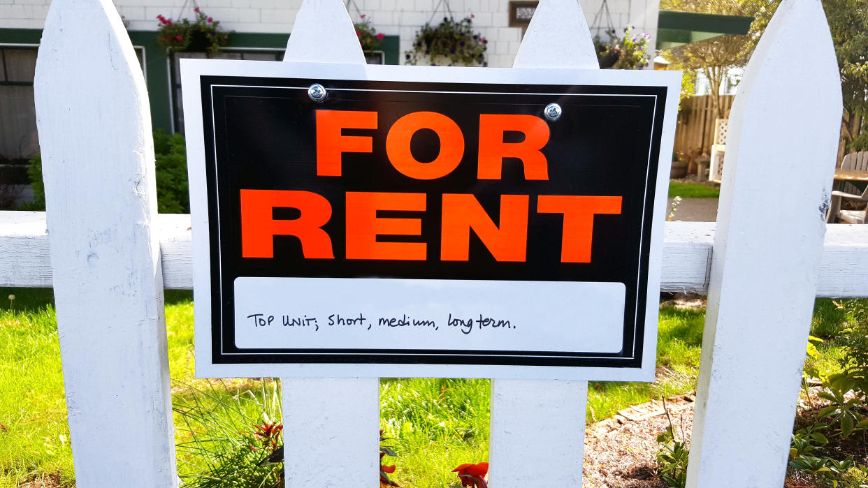 Close up of For Rent sign on white picket fence in front yard of house.