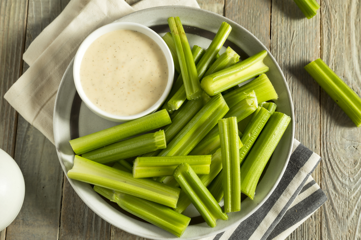 Top view of celery in a black ringed white ceramic bowl with a small white bowl of olive dip on a natural napkin, surrounded by a few celery sticks and a natural table