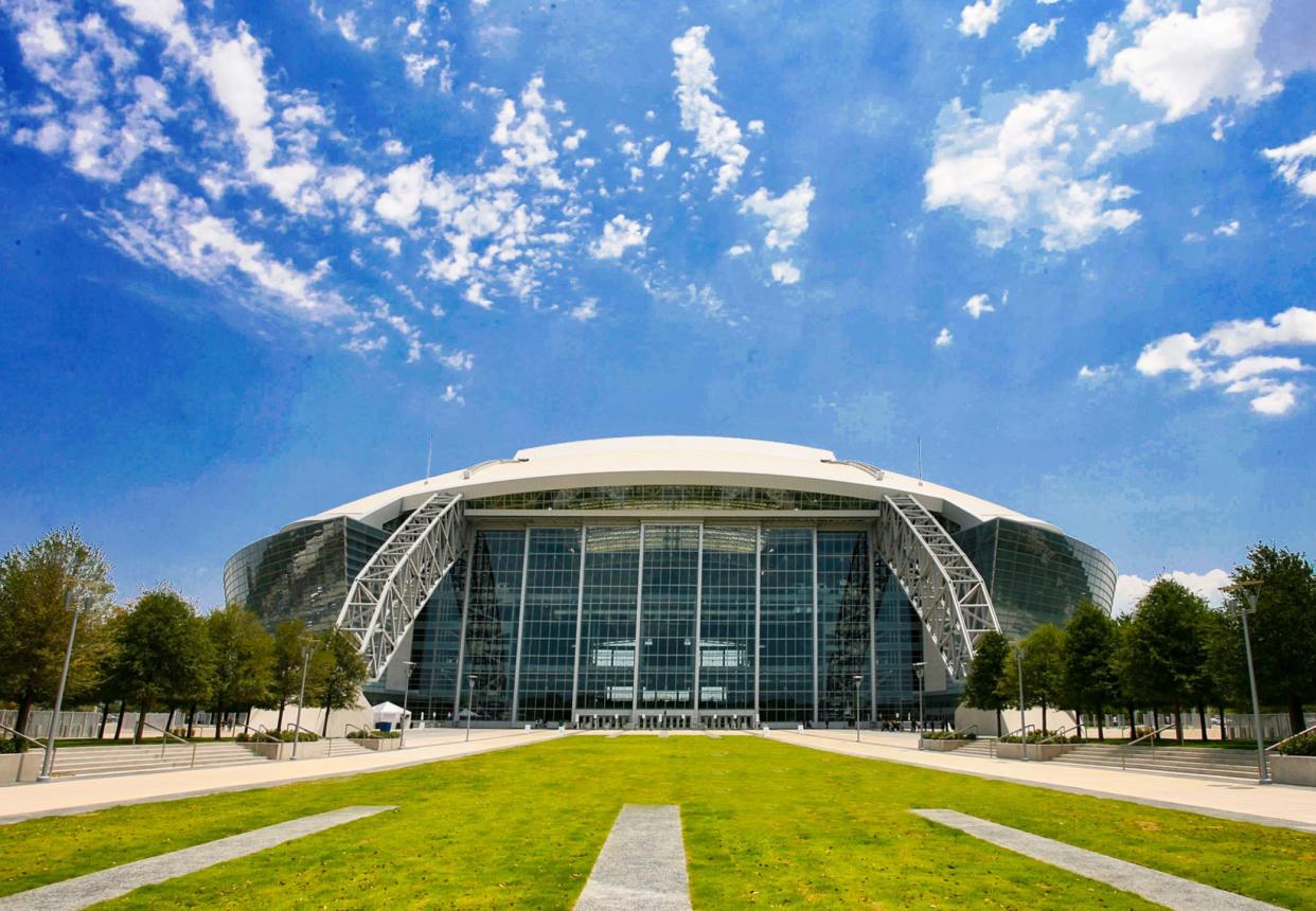 Dallas Cowboys, AT&T Stadium, Arlington, Texas, front exterior with grass in the foreground and against a dramatic blue sky with white clouds with trees and sidewalks on both sides