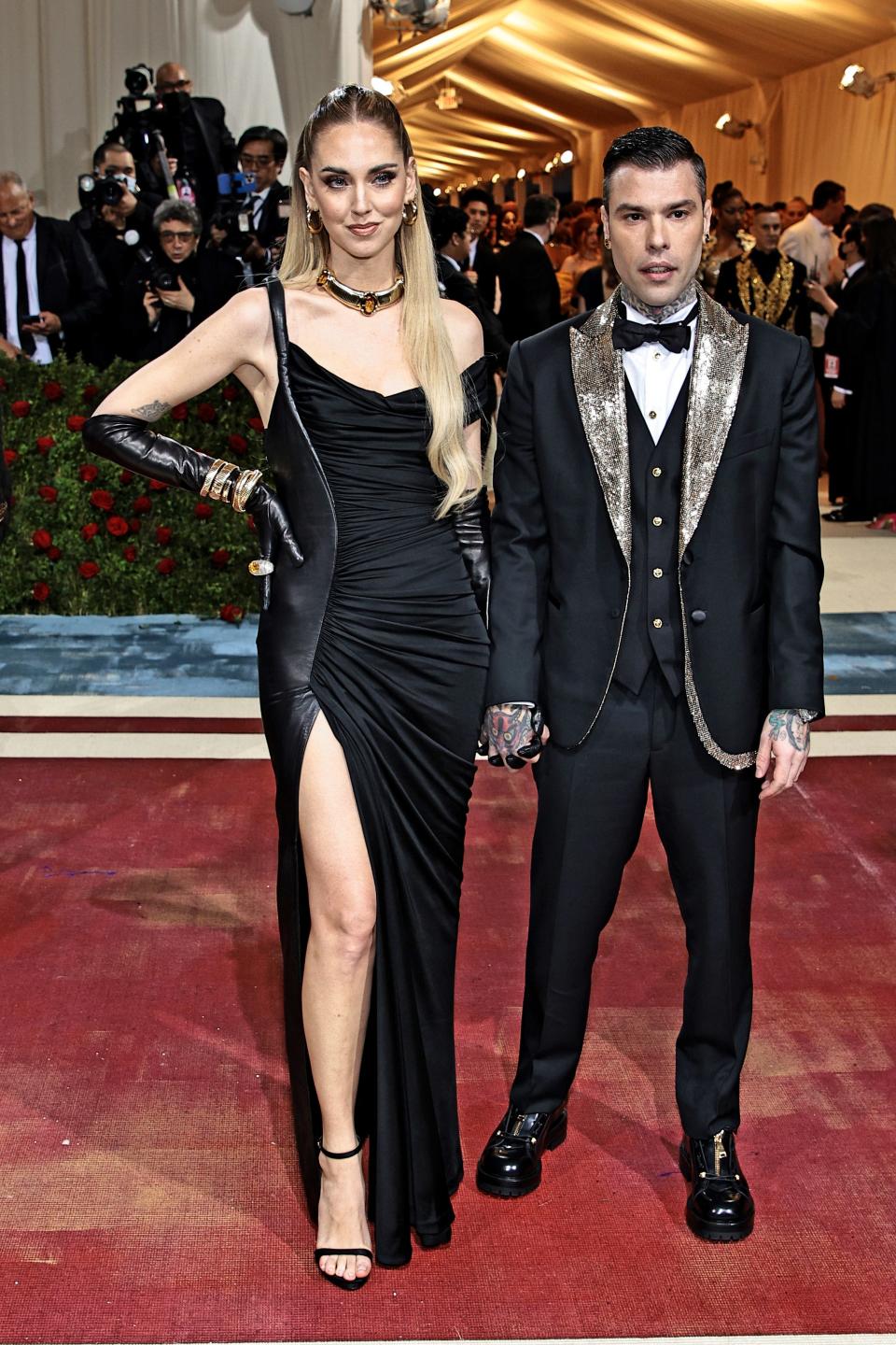 Chiara in a black gown, gloves, and heels and Fedez standing shorter in a black suit with metallic trim on a red carpet.
