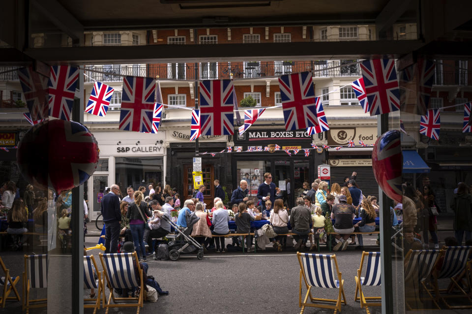 Varias personas comen su almuerzo en grandes mesas colocadas en la calle como parte de las celebraciones por la coronación del rey Carlos III, el domingo 7 de mayo de 2023, en Londres. (AP Foto/Emilio Morenatti)