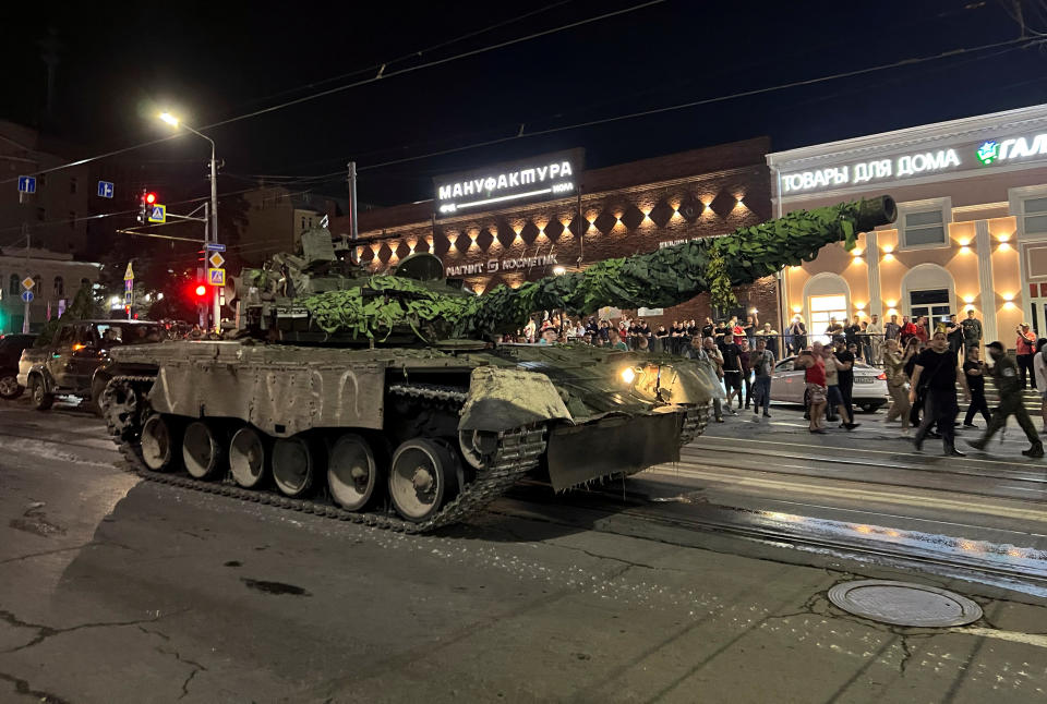 Dozens of onlookers stand near a tank in a road next to businesses with Russian language signs.