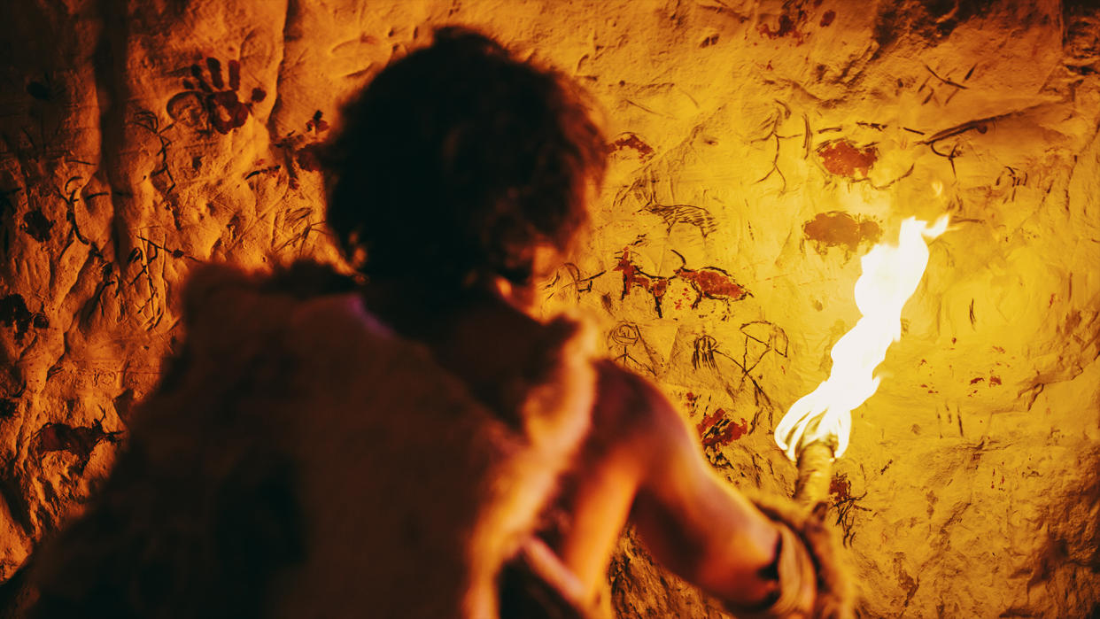  Primeval caveman wearing animal skin standing in his cave at night, holding torch with fire looking at drawings on the walls at night. Cave art with petroglyphs, rock paintings. 