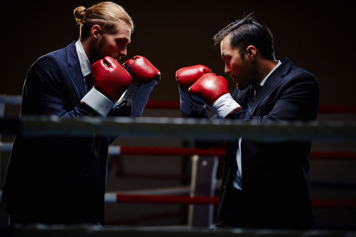 Two men in business suits facing each other in a boxing ring with their red gloved-fists up.