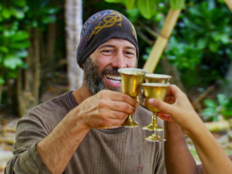 Tony Vlachos clinking a gold cup with the forest on the background on Survivor