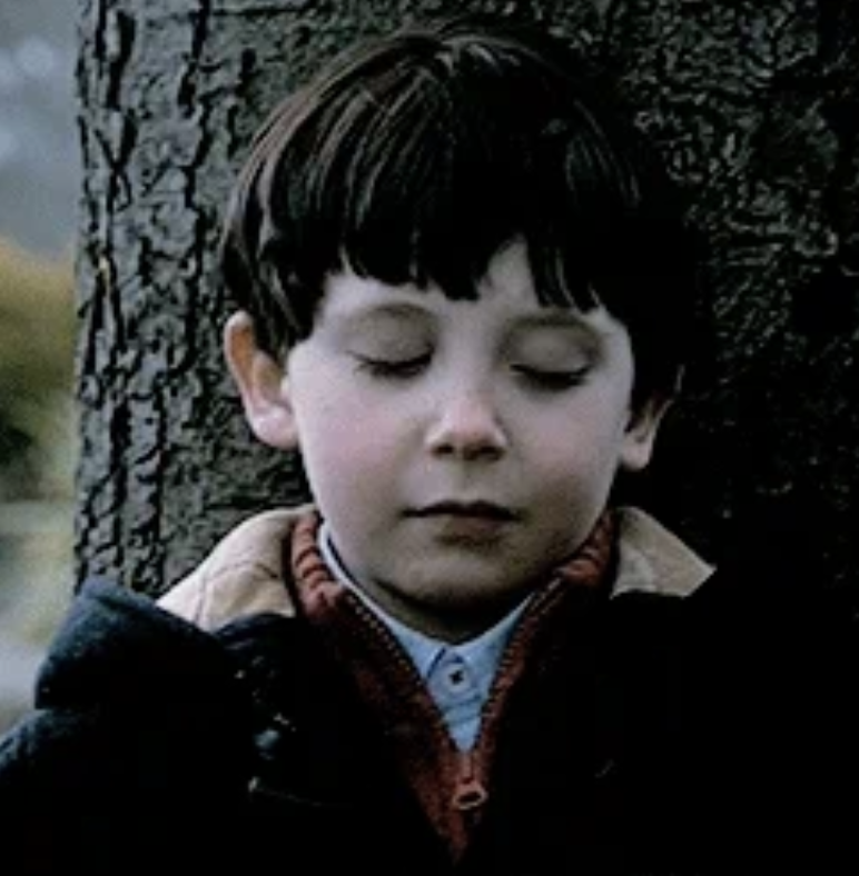 A young boy with closed eyes stands in front of a tree trunk