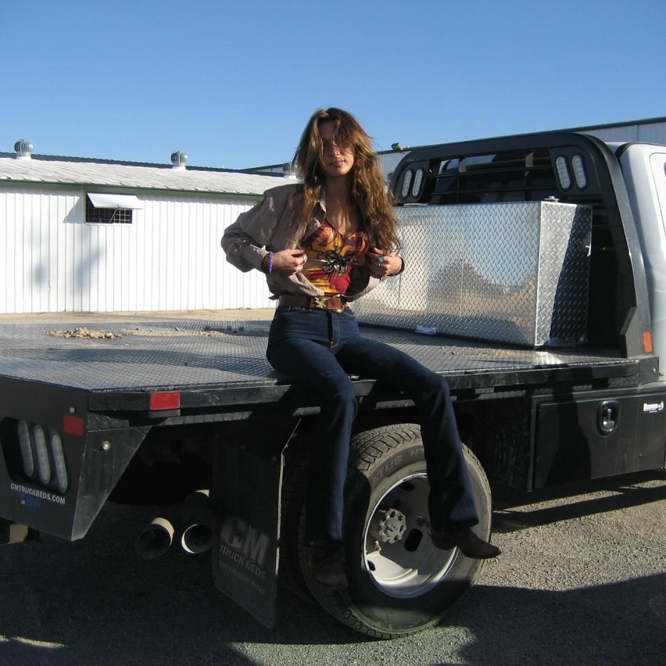 Bella Hadid sitting on a tractor wearing a colorful tank top with blue jeans and brown boots