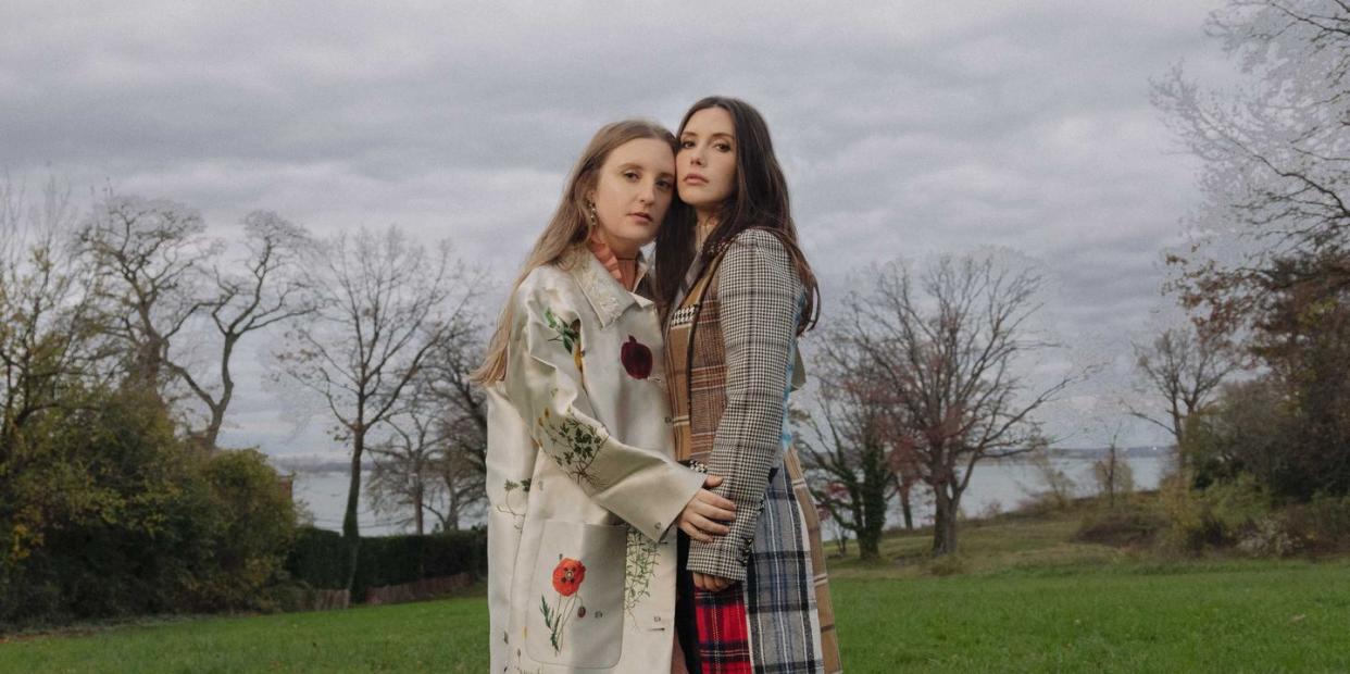 two sisters stand in a grassy field with a body of water in the background