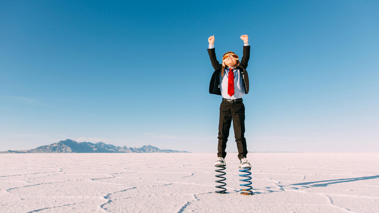 A young business boy dressed in business suit, flight cap and goggles stands on springs in the Utah desert.