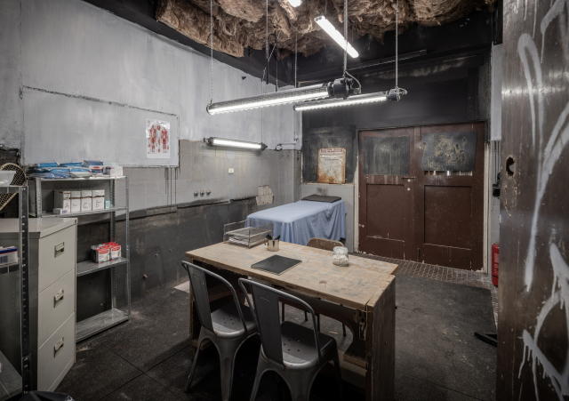 Basic medical room in disused building for SAS Australia, containing a medical examination bed, desk, filing cabinet and sparse shelving.