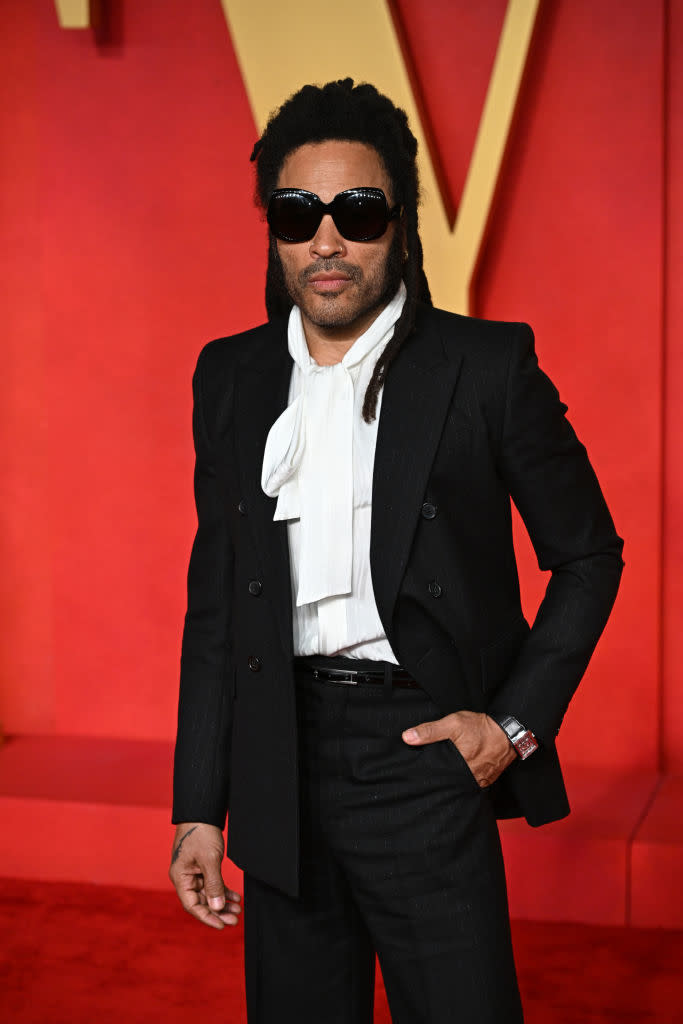 Lenny Kravitz in a suit with a ruffled white shirt, standing at an event with a red backdrop