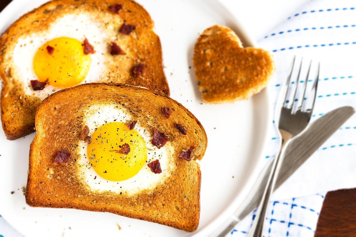 Focus on two slices of bread egg-in-a-hole, with heart-shaped holes, on a white plate with a fork and knife, on a blue dotted-lined white napkin