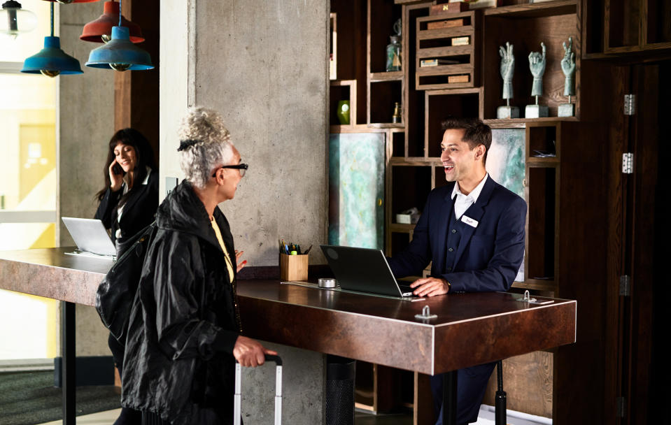 A person with gray hair in a bun is at a hotel reception desk, talking to a staff member in a suit. Another person is in the background on a phone call