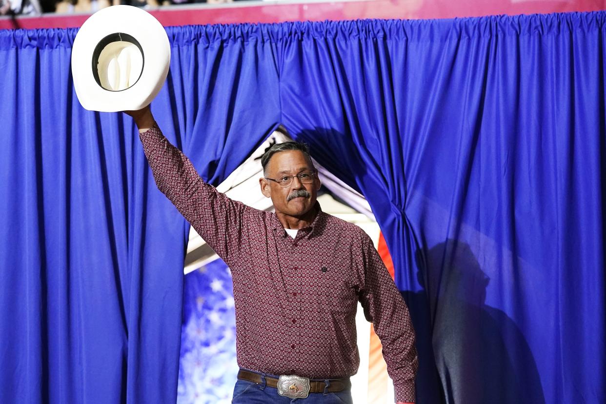 Mark Finchem, wearing a patterned button-down shirt and large belt buckle, stands in front of a blue curtain and waves with his hat.