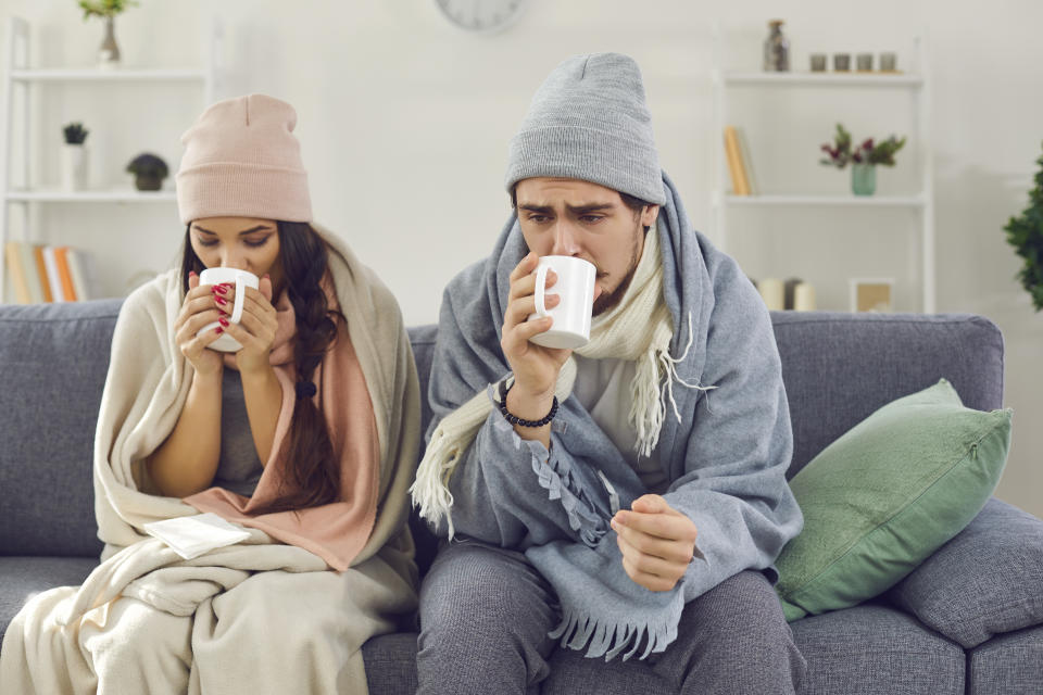 Sad sick young couple with norovirus wrapped in plaid sitting on sofa in living-room drinking hot tea trying to warm up. Freezing man and woman suffering from cold or flu fever or having trouble with central heating