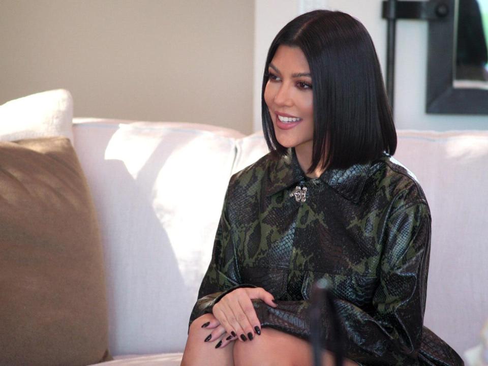 kourtney kardashian on the kardsahians, smiling and sitting on a white couch with her hands resting on her knees