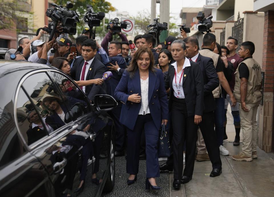 A woman in a suit, surrounded by a crowd of other people in suits, walks toward a shiny dark car