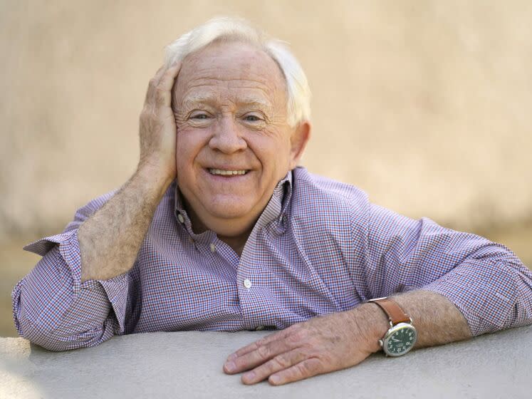 A man with white hair smiles while resting his cheek on his palm