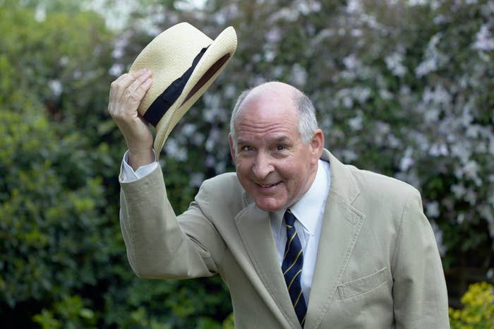 A person smiling, dressed in a light suit and striped tie, tips a straw hat outdoors