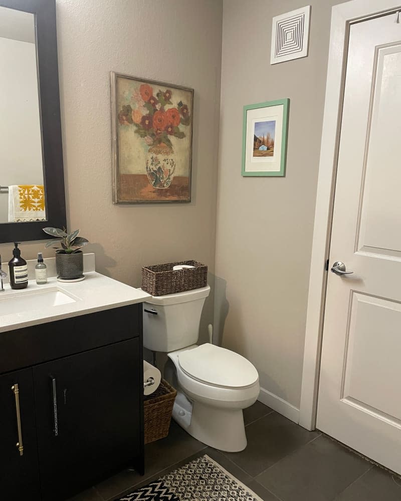 Floral art above toilet with basket in beige bathroom with dark wood framed mirror and cabinetry.