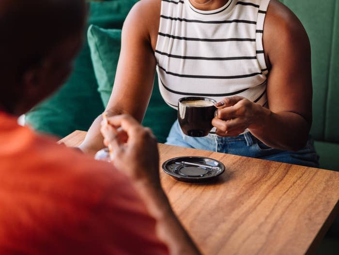 Woman in a striped sleeveless top with a ponytail smiling and holding a cup of coffee, sitting at a table with a man in a bright shirt