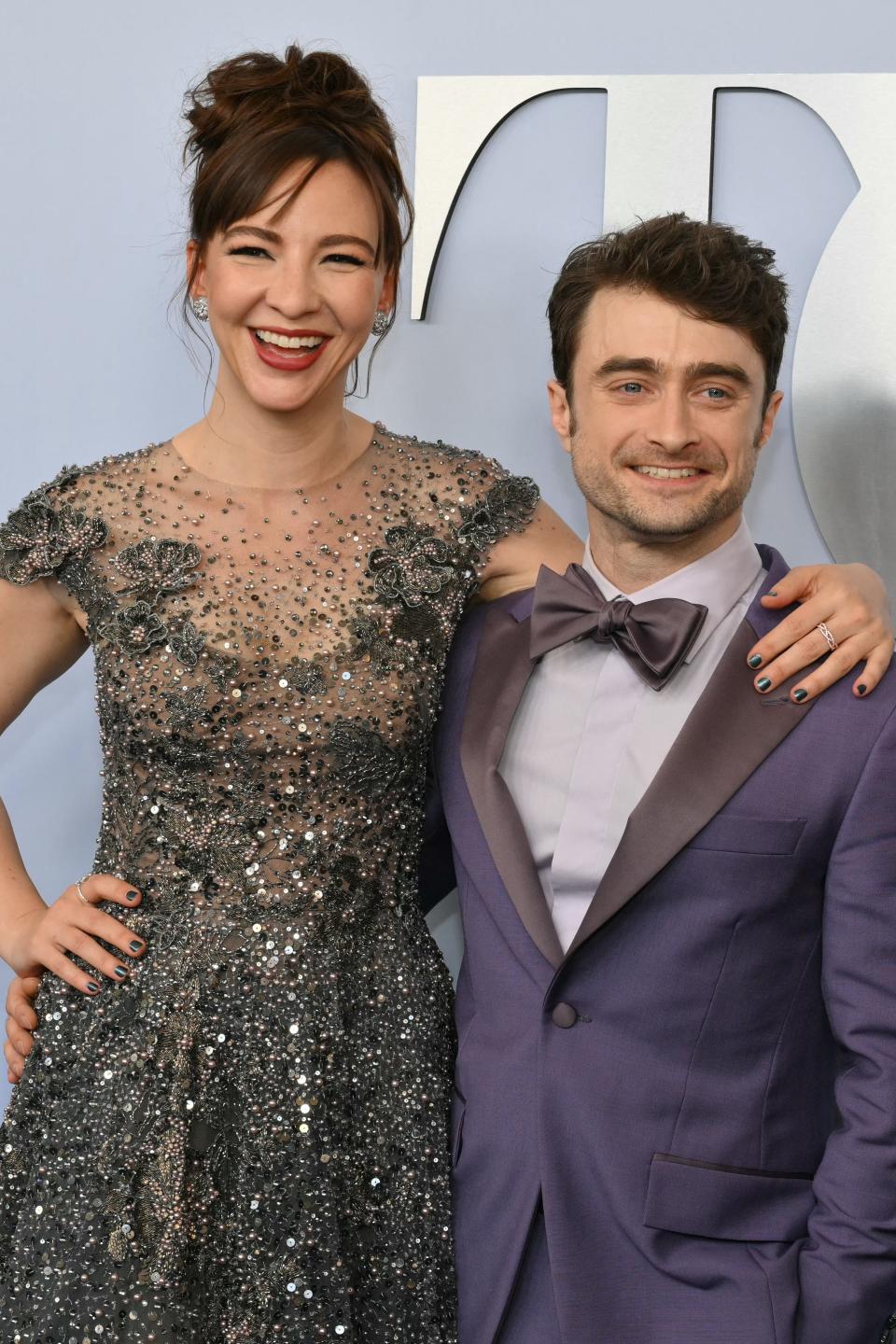 Daniel Radcliffe in a purple suit with a bow tie and Erin Darke in a sequined dress at a formal event, smiling for the camera