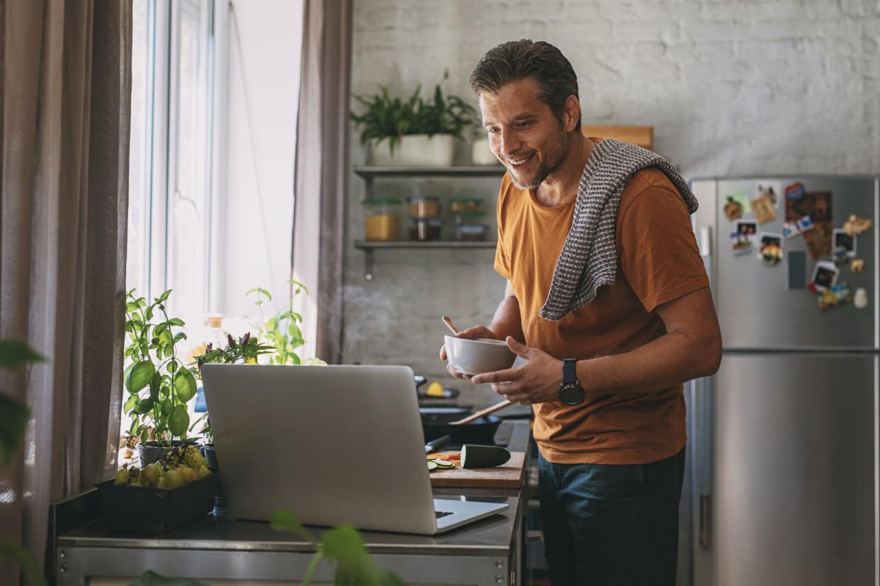 Handsome young happy Caucasian man holding a bowl, and making a meal while watching a cooking tutorial on a laptop on a kitchen counter.