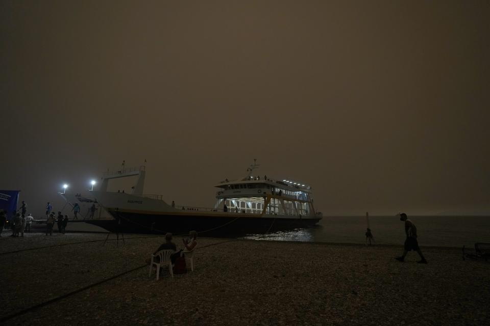 People gather on the beach as a ferry is docked for the evacuation during a wildfire at Pefki village on Evia island, about 189 kilometers (118 miles) north of Athens, Greece, Sunday, Aug. 8, 2021. Pillars of billowing smoke and ash are blocking out the sun above Greece's second-largest island as a days-old wildfire devours more pristine forests and triggers more evacuation alerts. (AP Photo/Petros Karadjias)