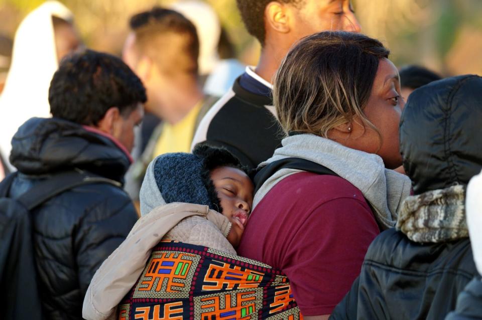 A mother, standing with a small group in the sunlight, carries a sleeping child on her back