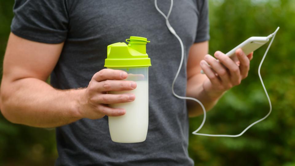 Nutritious protein cocktail in special shaker with a lot of milk for quenching the thirst and for taking a lot of useful vitamines. Male hand holds a smarthpone with headphones.