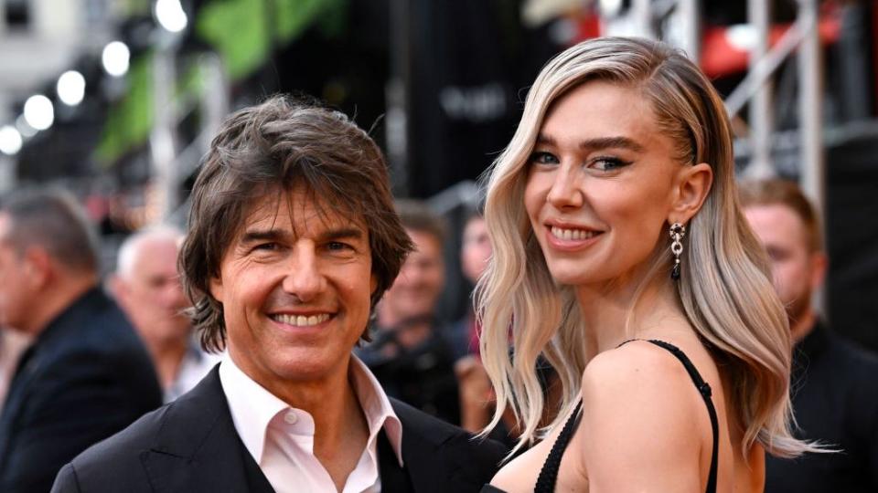 tom cruise, wearing a black suit jacket and pink shirt, smiling alongside vanessa kirby, who wears a black dress