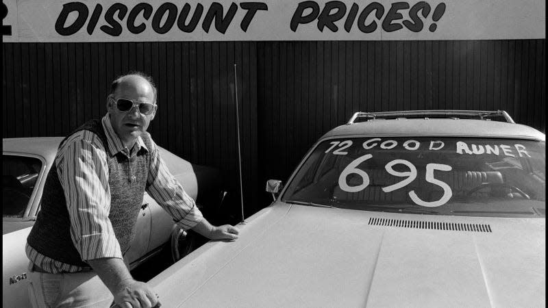 A black and white photo of a man leaning on a used car at a dealership. 
