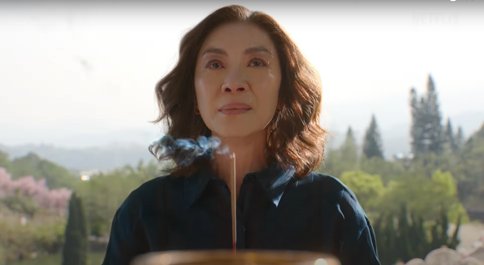 michelle yeoh, standing in front of trees and grasses in the distance, holding a cored incensed stick that's smoking at the top